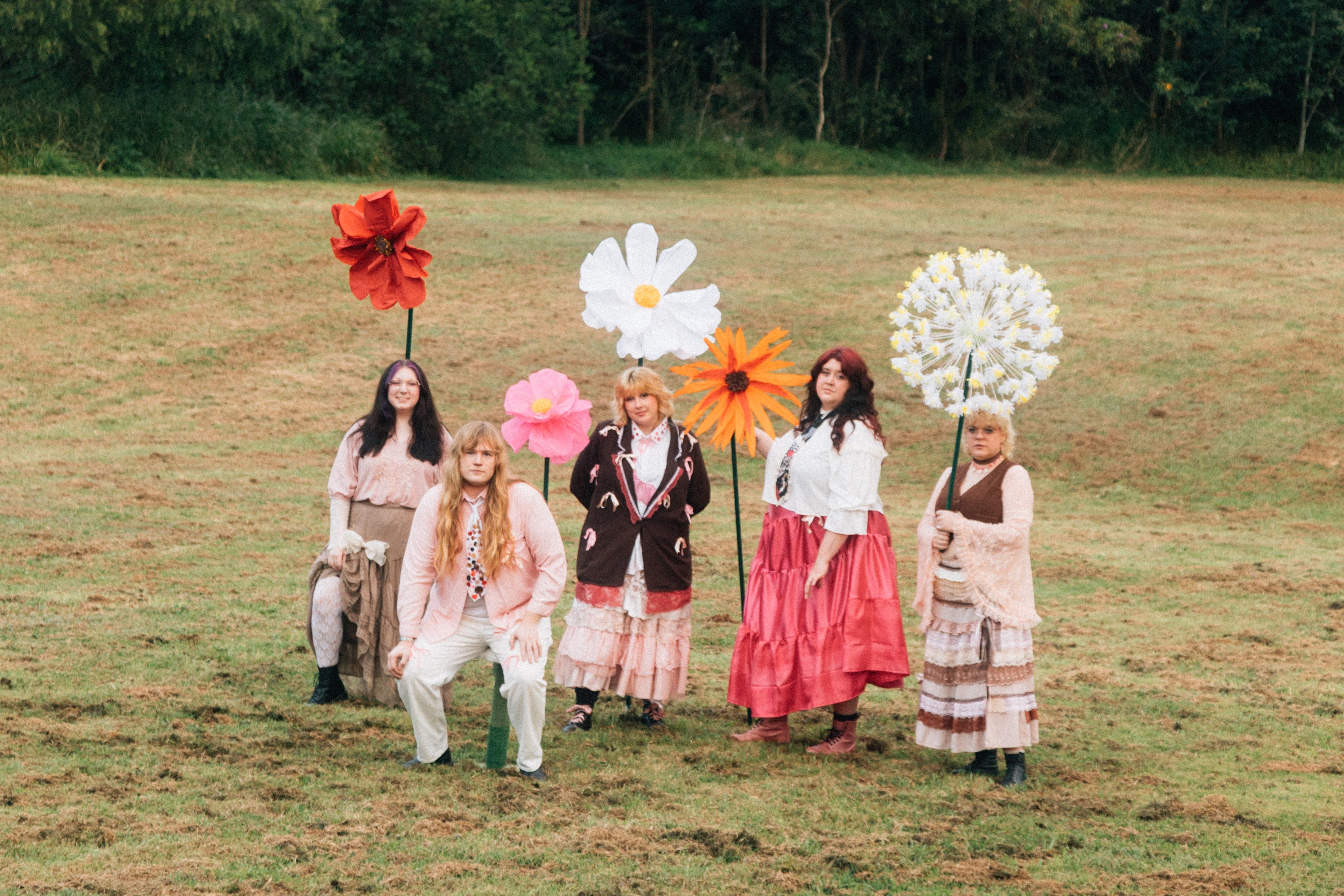 Five wormz in vintage-style clothing holding large flower props in a grassy field with trees in the background.